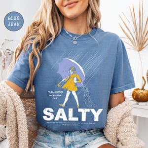 May include: Blue jean colored t-shirt with a graphic of a person under an umbrella, with the word "SALTY" in large white letters. The graphic includes the text "All blue flavors and you choose to be".