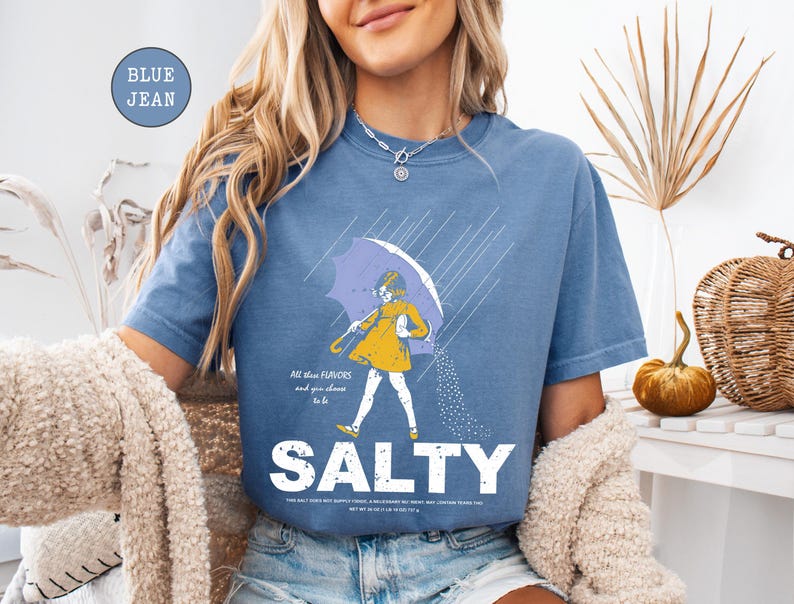 May include: Blue jean colored t-shirt with a graphic of a person under an umbrella, with the word "SALTY" in large white letters. The graphic includes the text "All blue flavors and you choose to be".