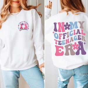 May include: White sweatshirt with a pink, blue, and purple graphic that says "In My Official Teenager Era" with flowers. The front of the sweatshirt has a smaller graphic with the number 13 and the name Olivia.