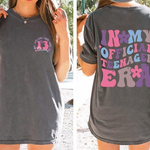 May include: A gray t-shirt with the text "In My Official Teenager Era" printed on the back in a retro font with pink, purple, and blue colors. The text is surrounded by flowers.