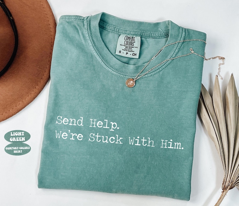 Pu&ograve; includere: T-shirt verde chiaro Comfort Colors con la scritta "Send Help. We're Stuck With Him." Una collana d'oro con un ciondolo &egrave; appoggiata sulla maglietta. Un cappello marrone e foglie di palma secche sono sullo sfondo.