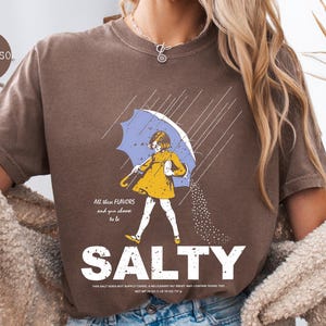 May include: Brown t-shirt with a graphic of a person under a blue umbrella, with the word "SALTY" in large white letters. The illustration includes the text "All these flavors and you choose to be".