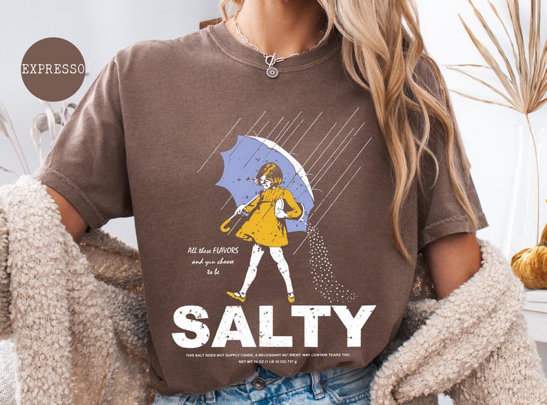May include: Brown t-shirt with a graphic of a person under a blue umbrella, with the word "SALTY" in large white letters. The illustration includes the text "All these flavors and you choose to be".