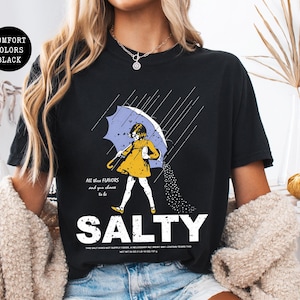 May include: Black t-shirt with a graphic of a person under an umbrella, with the word "SALTY" in large white letters. The person is wearing a yellow dress and holding a light blue umbrella. The text "All these flavors and you choose to be" is above the person.
