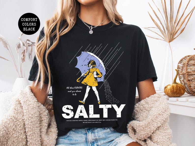 May include: Black t-shirt with a graphic of a person under an umbrella, with the word "SALTY" in large white letters. The person is wearing a yellow dress and holding a light blue umbrella. The text "All these flavors and you choose to be" is above the person.