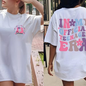 May include: White t-shirt with a pink and blue graphic that says "Official Teenager Era" with flowers on the back. The front of the shirt has a pink and blue graphic that says "Official Teenager Era" with flowers and the name "Olivia" underneath.