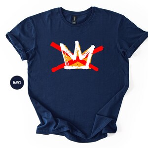 May include: Navy blue t-shirt featuring a graphic crown design in white, orange, and yellow, crossed by two red lines. The shirt has short sleeves and a crew neck. The word "NAVY" is in a navy circle.