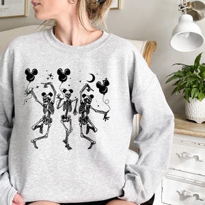 May include: A light gray sweatshirt featuring a black graphic of three dancing skeletons holding Mickey Mouse-shaped balloons. The skeletons are depicted with detailed rib cages and limbs, with stars and a crescent moon in the background.