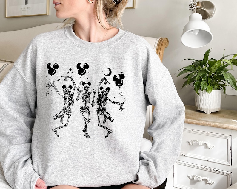 May include: A light gray sweatshirt featuring a black graphic of three dancing skeletons holding Mickey Mouse-shaped balloons. The skeletons are depicted with detailed rib cages and limbs, with stars and a crescent moon in the background.