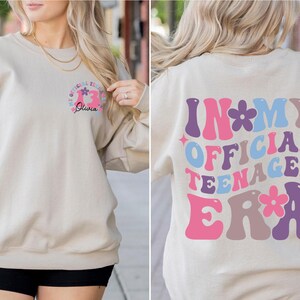 May include: A beige sweatshirt with a pink, blue, and purple graphic that says "In My Official Teenager Era" with flowers. The front of the sweatshirt has a smaller graphic with a pink number 13 and the name "Olivia" in black.