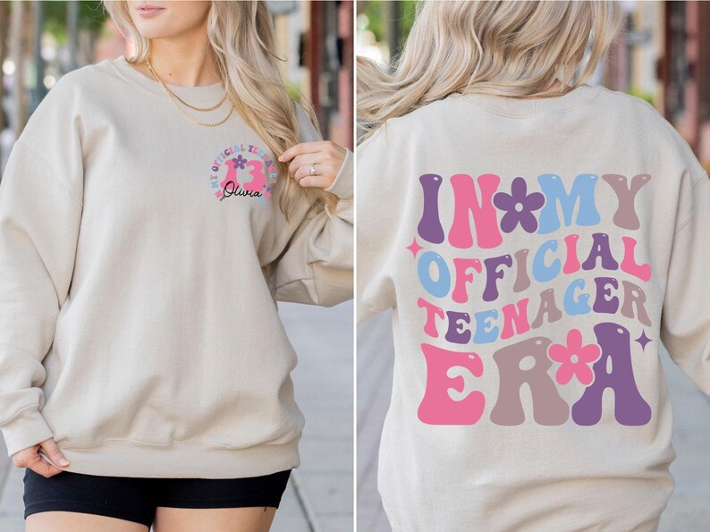 May include: A beige sweatshirt with a pink, blue, and purple graphic that says "In My Official Teenager Era" with flowers. The front of the sweatshirt has a smaller graphic with a pink number 13 and the name "Olivia" in black.