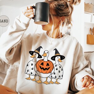 Peut inclure: Sweat-shirt cr&egrave;me avec un motif d'Halloween. Le dessin repr&eacute;sente trois canards, deux portant des chapeaux de sorci&egrave;re et des capes fant&ocirc;mes, et un avec un corps de citrouille. Les canards ont des becs et des pattes orange.