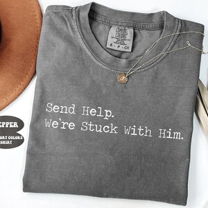 Pu&ograve; includere: Una maglietta grigia Comfort Colors con la scritta "Send Help. We're Stuck With Him." Una collana d'argento con un piccolo ciondolo &egrave; appoggiata sulla maglietta. La maglietta &egrave; piegata e appoggiata su una superficie bianca.