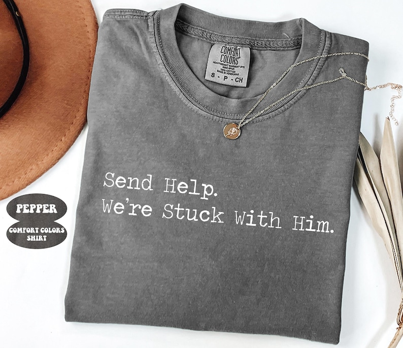 Pu&ograve; includere: Una maglietta grigia Comfort Colors con la scritta "Send Help. We're Stuck With Him." Una collana d'argento con un piccolo ciondolo &egrave; appoggiata sulla maglietta. La maglietta &egrave; piegata e appoggiata su una superficie bianca.