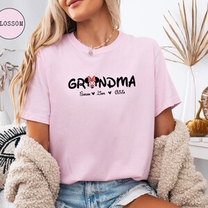Op de afbeelding: Roze T-shirt met een zwarte afbeelding waarop "Grandma" staat met een Minnie Mouse hoofd in het midden. De tekst "Sarah Liam Olivia" staat onder de afbeelding.