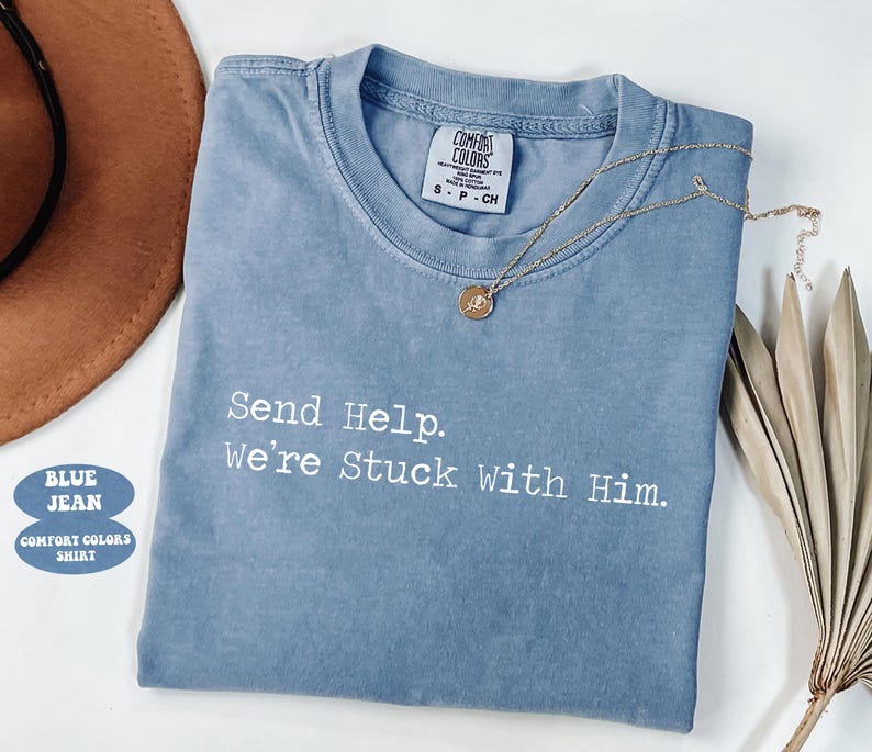 Pu&ograve; includere: Una maglietta Comfort Colors blu jeans con la scritta "Send Help. We're Stuck With Him." Una collana d'oro con un ciondolo &egrave; drappeggiata sulla maglietta. Un cappello marrone e foglie di palma secche sono sullo sfondo.