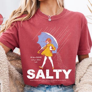 May include: Crimson-colored t-shirt with a graphic of a person under a blue umbrella, wearing a yellow dress, and the word "SALTY" in large white letters. The text "All these flavors and you choose to be" is above the graphic.