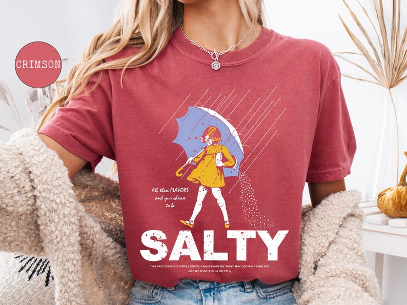 May include: Crimson-colored t-shirt with a graphic of a person under a blue umbrella, wearing a yellow dress, and the word "SALTY" in large white letters. The text "All these flavors and you choose to be" is above the graphic.