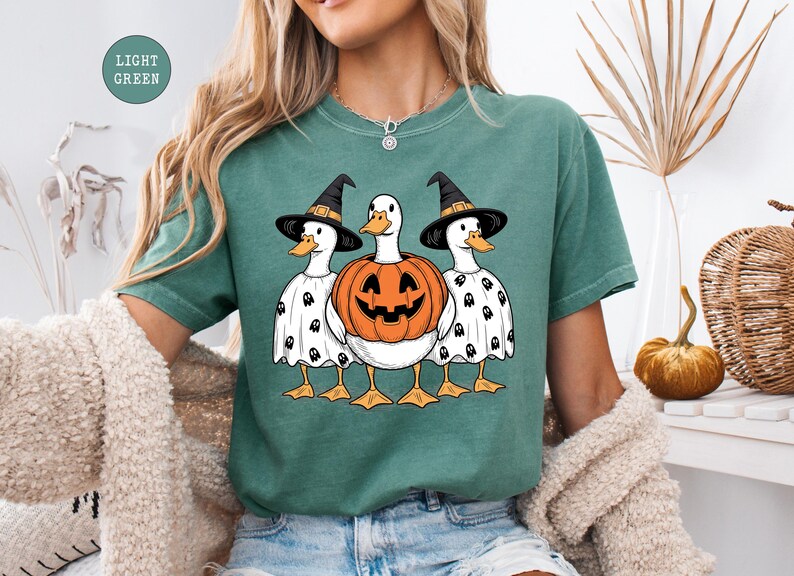 Peut inclure: T-shirt vert clair avec un motif d'Halloween. Le motif repr&eacute;sente trois canards de dessin anim&eacute; portant des chapeaux de sorci&egrave;re et des capes de fant&ocirc;mes. Le canard du milieu est une citrouille sculpt&eacute;e. Les canards ont des becs et des pattes oranges.
