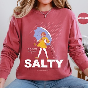 May include: A maroon long-sleeve sweatshirt with a graphic of a person under an umbrella, with the word "SALTY" in large white letters. The graphic includes the text "All these flavors and you choose to be".