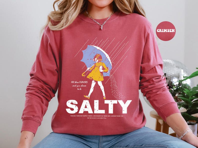 May include: A maroon long-sleeve sweatshirt with a graphic of a person under an umbrella, with the word "SALTY" in large white letters. The graphic includes the text "All these flavors and you choose to be".