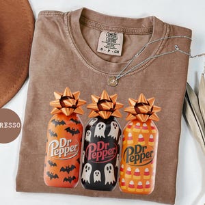May include: Espresso-colored t-shirt with a crew neck, featuring three decorative Dr. Pepper bottles with Halloween-themed designs. The bottles have orange, black, and yellow patterns with bow-shaped toppers. A silver necklace with a pendant is also visible.
