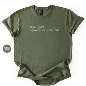 Pu&ograve; includere: T-shirt verde oliva con la scritta bianca "Send Help. We're Stuck with Him.". La maglietta ha un girocollo e maniche arrotolate. Una piccola etichetta circolare sul lato sinistro recita "MILITARY GREEN".