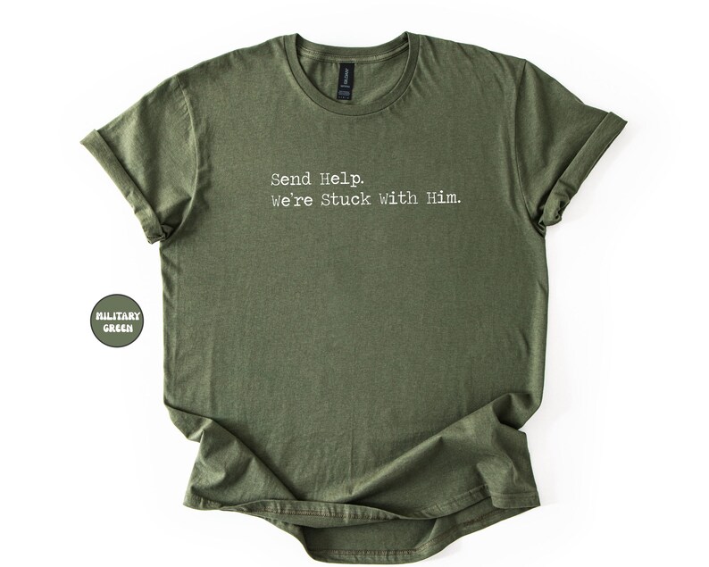 Pu&ograve; includere: T-shirt verde oliva con la scritta bianca "Send Help. We're Stuck with Him.". La maglietta ha un girocollo e maniche arrotolate. Una piccola etichetta circolare sul lato sinistro recita "MILITARY GREEN".