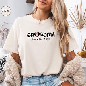 Op de afbeelding: Een wit T-shirt met de tekst "Grandma" in zwart met een rood en wit Minnie Mouse hoofd-ontwerp. De tekst "Sarah, Liam, Olivia" is onder het ontwerp gedrukt.