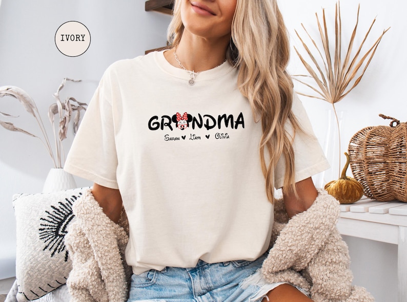 Op de afbeelding: Een wit T-shirt met de tekst "Grandma" in zwart met een rood en wit Minnie Mouse hoofd-ontwerp. De tekst "Sarah, Liam, Olivia" is onder het ontwerp gedrukt.