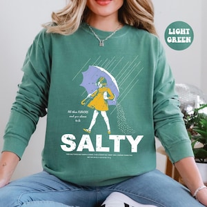 May include: Light green sweatshirt with a graphic of a person under an umbrella, with the word "SALTY" in large white letters. The person is wearing a yellow dress and holding a purple umbrella. Text on the shirt reads "All the flavors and you choose to be."