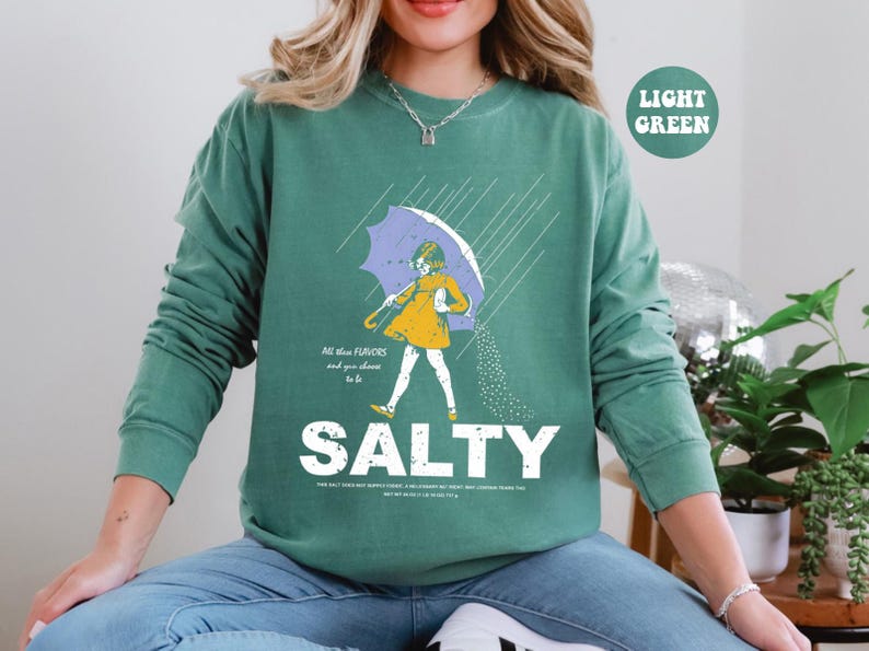 May include: Light green sweatshirt with a graphic of a person under an umbrella, with the word "SALTY" in large white letters. The person is wearing a yellow dress and holding a purple umbrella. Text on the shirt reads "All the flavors and you choose to be."