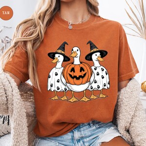 Peut inclure: T-shirt orange avec un motif d'Halloween. Trois canards sont v&ecirc;tus de chapeaux de sorci&egrave;re et de costumes de fant&ocirc;mes, avec une citrouille sculpt&eacute;e au centre. Les canards ont des becs et des pieds jaunes. Le t-shirt est de couleur orange unie.
