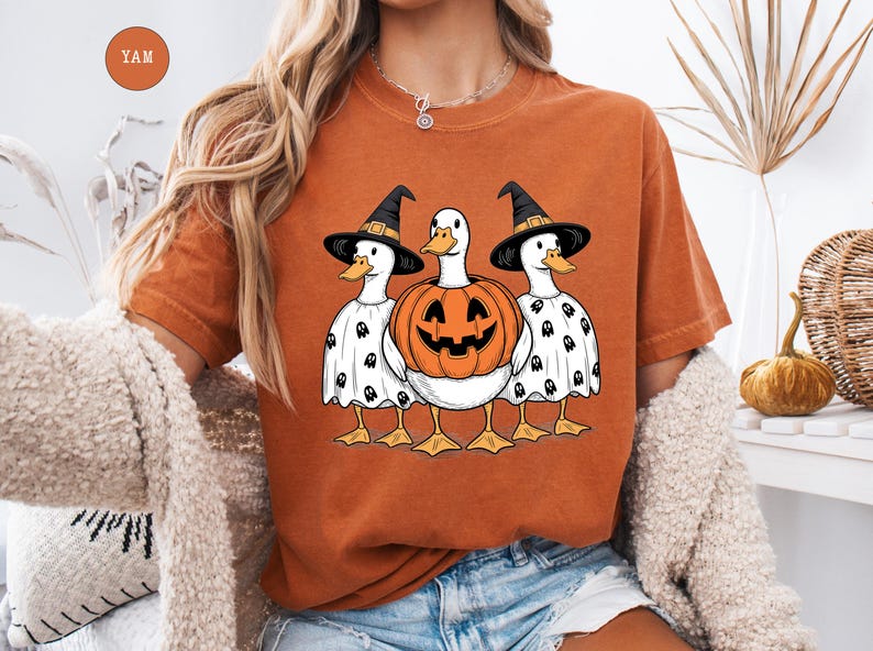 Peut inclure: T-shirt orange avec un motif d'Halloween. Trois canards sont v&ecirc;tus de chapeaux de sorci&egrave;re et de costumes de fant&ocirc;mes, avec une citrouille sculpt&eacute;e au centre. Les canards ont des becs et des pieds jaunes. Le t-shirt est de couleur orange unie.