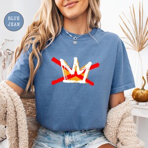 May include: Blue jean-colored t-shirt featuring a graphic of a crown with red and yellow accents. The crown design is crossed out with two red lines. The shirt is a short-sleeved, casual style.