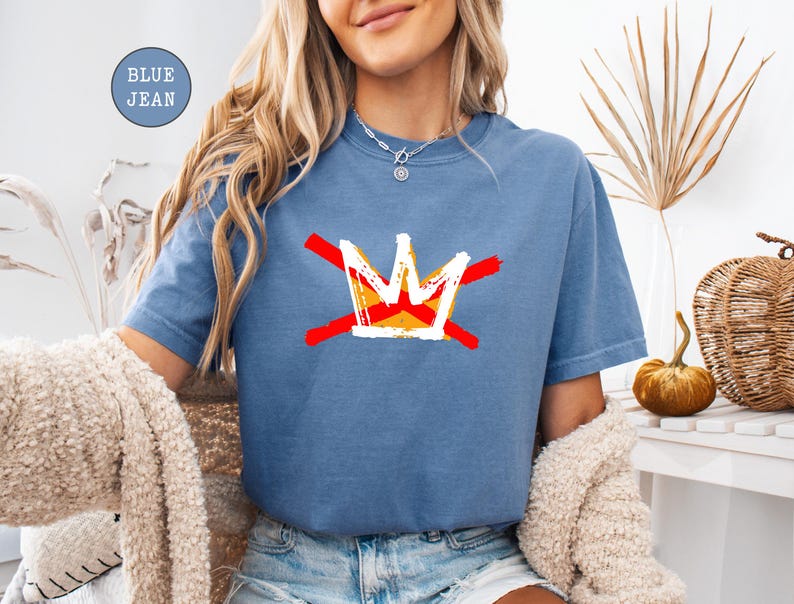 May include: Blue jean-colored t-shirt featuring a graphic of a crown with red and yellow accents. The crown design is crossed out with two red lines. The shirt is a short-sleeved, casual style.