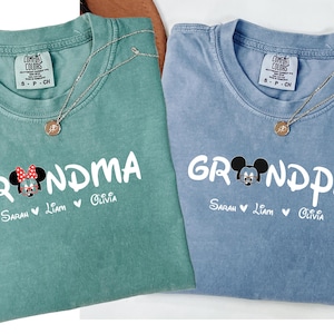 Op de afbeelding: Twee lichtblauwe en groene t-shirts met een Mickey Mouse en Minnie Mouse afbeelding. De tekst op de shirts luidt "Grandma" en "Grandpa" met de namen Sarah, Liam en Olivia eronder.