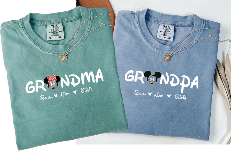 Op de afbeelding: Twee lichtblauwe en groene t-shirts met een Mickey Mouse en Minnie Mouse afbeelding. De tekst op de shirts luidt "Grandma" en "Grandpa" met de namen Sarah, Liam en Olivia eronder.