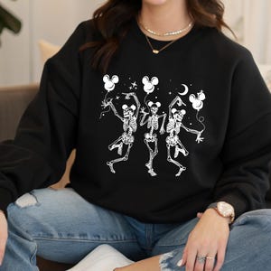 May include: Black sweatshirt featuring a white graphic of three dancing skeletons holding Mickey Mouse-shaped balloons. The skeletons are in a playful pose, with stars and a crescent moon in the background. A fun, Halloween-themed design.