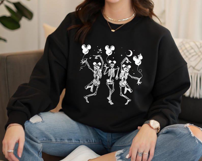 May include: Black sweatshirt featuring a white graphic of three dancing skeletons holding Mickey Mouse-shaped balloons. The skeletons are in a playful pose, with stars and a crescent moon in the background. A fun, Halloween-themed design.