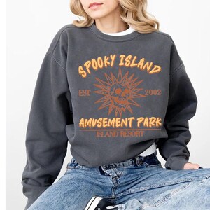 A woman wearing a grey sweatshirt with orange and white text and graphics, including a skull and sun design, sitting on a chair.