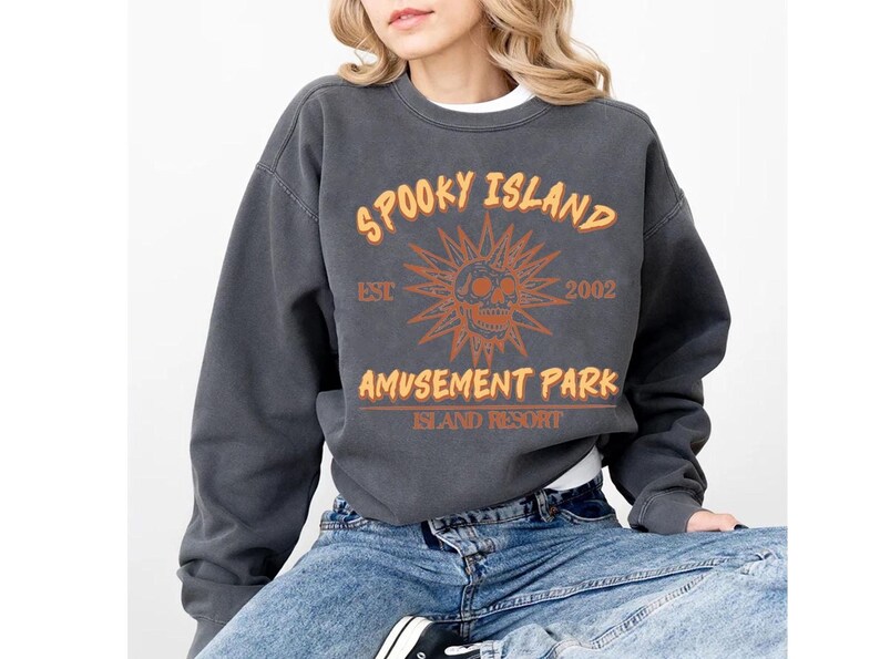 A woman wearing a grey sweatshirt with orange and white text and graphics, including a skull and sun design, sitting on a chair.