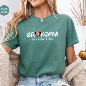 Op de afbeelding: Een lichtgroen T-shirt met het woord "Grandma" in witte letters en een cartoonafbeelding van Minnie Mouse. Het shirt heeft ook de namen "Sarah, Liam, Olivia" gedrukt onder het woord "Grandma".