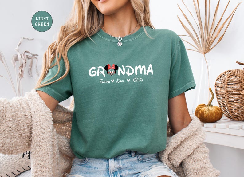 Op de afbeelding: Een lichtgroen T-shirt met het woord "Grandma" in witte letters en een cartoonafbeelding van Minnie Mouse. Het shirt heeft ook de namen "Sarah, Liam, Olivia" gedrukt onder het woord "Grandma".