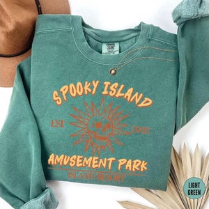 A green sweatshirt with orange text and a skull design, along with a brown hat and some leaves, is displayed on a white background.