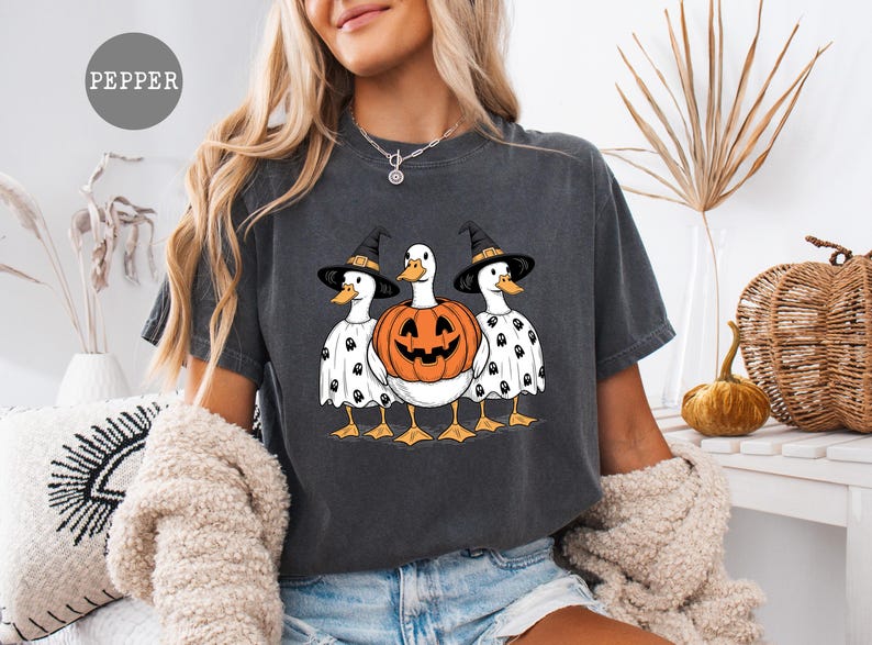 Peut inclure: T-shirt gris fonc&eacute; avec un motif d'Halloween. Le graphisme repr&eacute;sente trois canards, deux portant des chapeaux de sorci&egrave;re et des costumes de fant&ocirc;mes, et un en citrouille. Les canards sont regroup&eacute;s, cr&eacute;ant une image festive et fantaisiste.