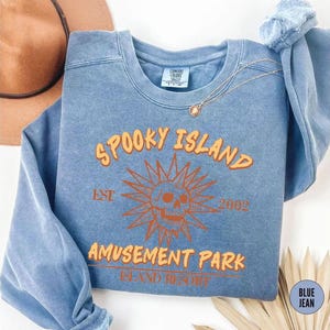A blue sweatshirt with orange text and a skull graphic, along with a necklace and a hat, is displayed on a white surface.