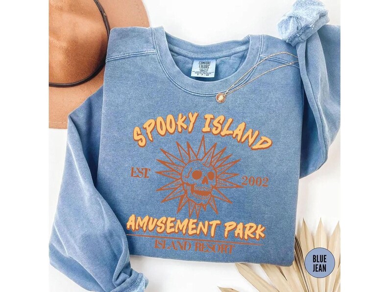 A blue sweatshirt with orange text and a skull graphic, along with a necklace and a hat, is displayed on a white surface.