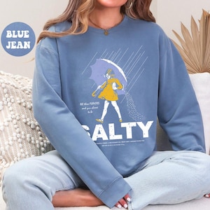 May include: Blue jean sweatshirt with a graphic of a person under an umbrella, pouring salt. The word "SALTY" is printed in large white letters below the graphic. The text "All the flavors and you choose to be" is above the word "SALTY".