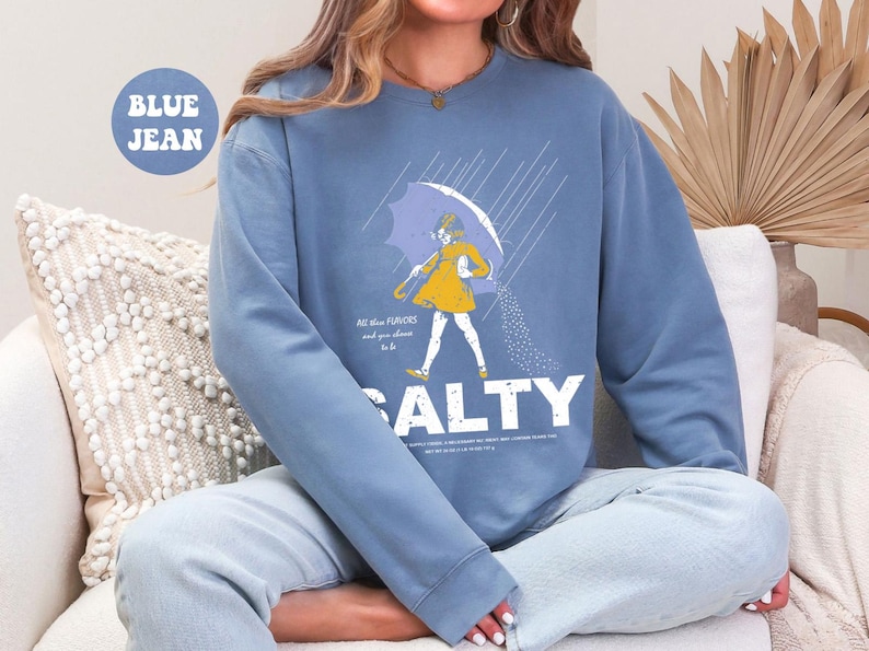 May include: Blue jean sweatshirt with a graphic of a person under an umbrella, pouring salt. The word "SALTY" is printed in large white letters below the graphic. The text "All the flavors and you choose to be" is above the word "SALTY".